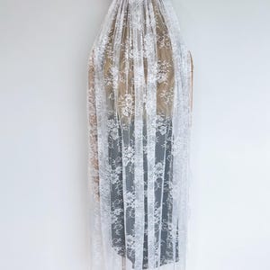 Op de afbeelding: Een lange witte kanten sluier met een gedetailleerd bloemenpatroon over de gehele stof. De sluier is aan de bovenkant geplooid en vastgezet met witte bloemaccenten, waardoor deze in een transparant, gelaagd effect over een donker kledingstuk valt.