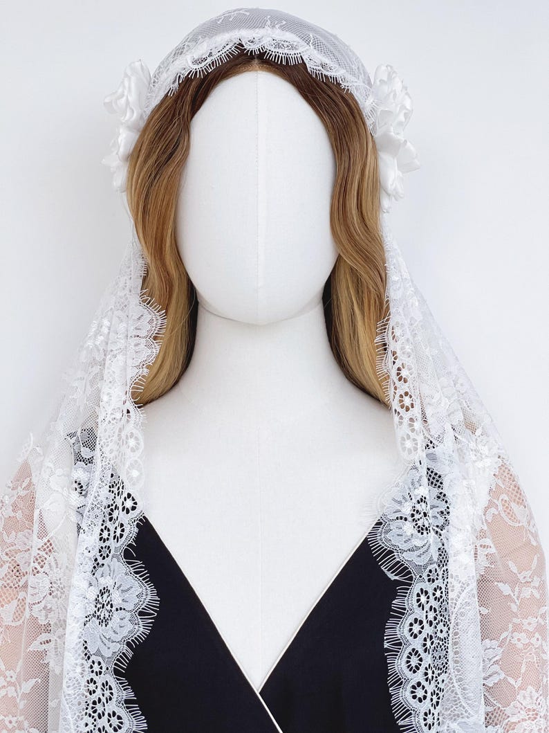 Op de afbeelding: Een witte kanten mantillasluier met ingewikkelde bloemmotieven en geschulpte randen, gedrapeerd over een etalagepophoofd met lang golvend lichtbruin haar. Twee witte stoffen bloemen zijn aan de zijkanten van het hoofddeksel bevestigd.