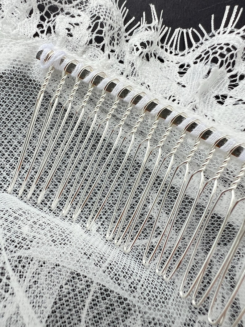 Op de afbeelding: Een zilverkleurige haarkam met een rij dunne, metalen tanden met gedraaide draaddetails aan de basis. De kam is bevestigd aan een stuk witte kanten stof met een delicaat, ingewikkeld bloemenpatroon en geschulpte randen.