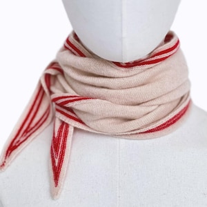 May include: A beige knit scarf featuring vibrant red striped borders and pointed ends. The scarf is wrapped multiple times around the neck, showcasing a soft, textured fabric with a distinct chevron-like pattern at the tapered edges.