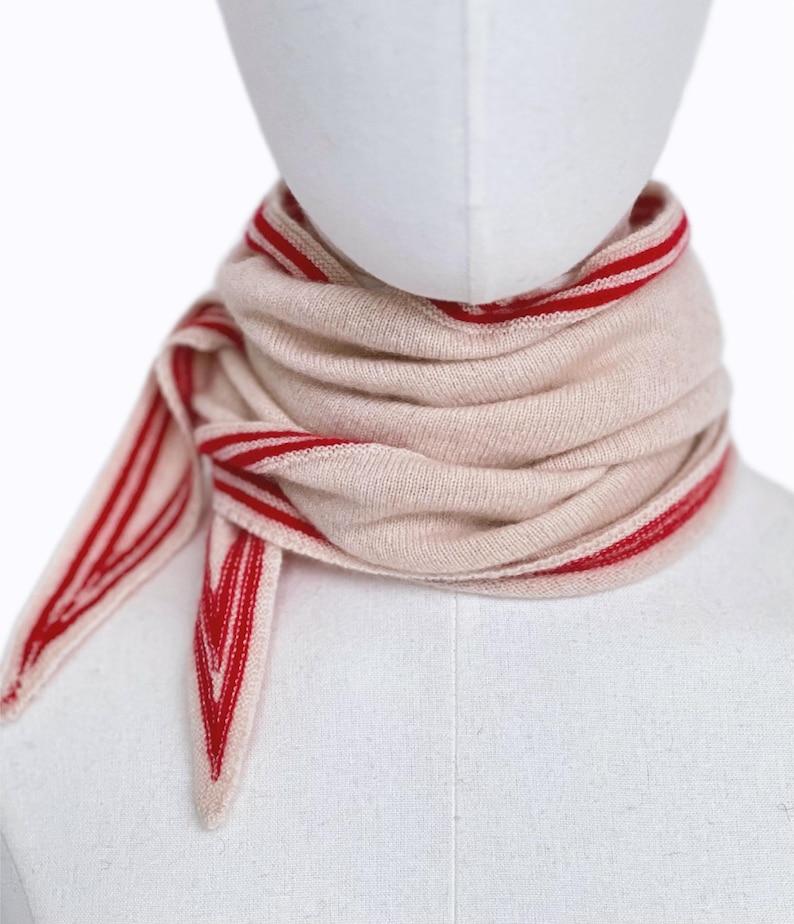 May include: A beige knit scarf featuring vibrant red striped borders and pointed ends. The scarf is wrapped multiple times around the neck, showcasing a soft, textured fabric with a distinct chevron-like pattern at the tapered edges.