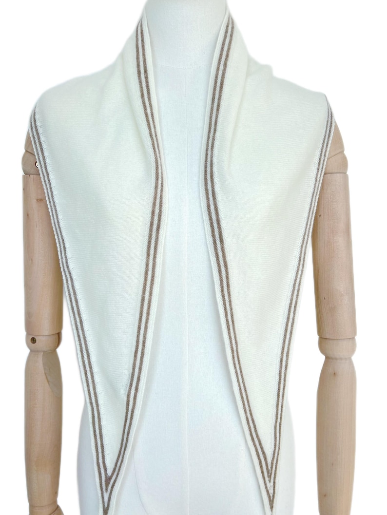 May include: A triangular scarf in off-white knit fabric featuring a double-line border in a light brown tone. The scarf is draped symmetrically, showcasing the clean edges and the consistent color contrast between the cream base and the brown trim.