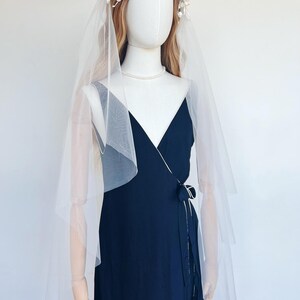 May include: A mannequin wearing a navy blue wrap dress, a sheer white veil, a floral crown, and a pearl necklace. The dress has a tie at the waist. The veil drapes over the shoulders. The floral crown is white.