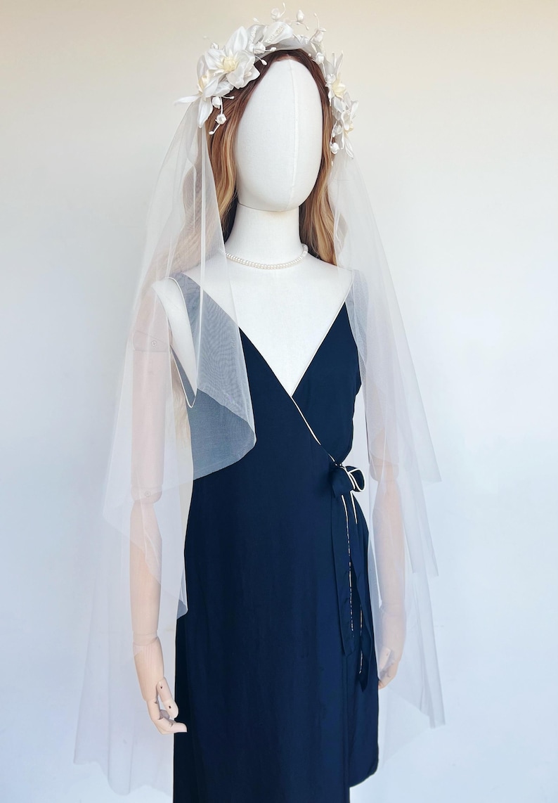 May include: A mannequin wearing a navy blue wrap dress, a sheer white veil, a floral crown, and a pearl necklace. The dress has a tie at the waist. The veil drapes over the shoulders. The floral crown is white.