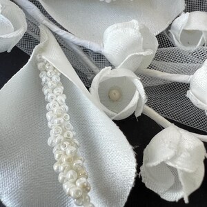 May include: Close-up of a white floral arrangement featuring fabric petals, beaded accents, and small, closed rosebuds. The arrangement includes delicate white stems and a mesh backing, creating a textured and elegant design.