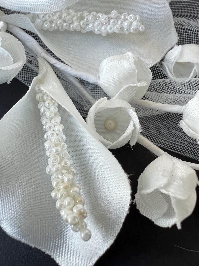May include: Close-up of a white floral arrangement featuring fabric petals, beaded accents, and small, closed rosebuds. The arrangement includes delicate white stems and a mesh backing, creating a textured and elegant design.