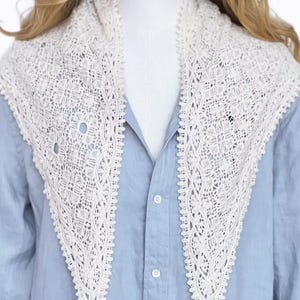 May include: A cream-coloured lace collar with an intricate openwork pattern. The collar, triangular in shape, is worn over a light blue shirt. The lace has a delicate, textured appearance, and the edges of the collar are scalloped.