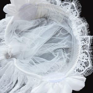 Op de afbeelding: Een wit bruidshoofddeksel met twee satijnen bloemen bevestigd aan een flexibele band. Het accessoire bevat een doorschijnende tule sluier en een delicate kanten rand langs de zijkanten. Twee zilverkleurige metalen haarkammen zijn aan de onderzijde bevestigd voor een stevige grip op de gelaagde stof en de bloemaccenten.