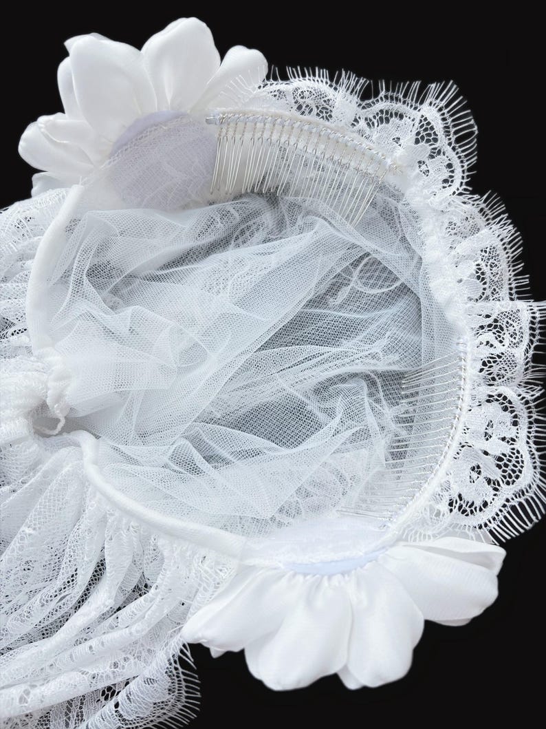 Op de afbeelding: Een wit bruidshoofddeksel met twee satijnen bloemen bevestigd aan een flexibele band. Het accessoire bevat een doorschijnende tule sluier en een delicate kanten rand langs de zijkanten. Twee zilverkleurige metalen haarkammen zijn aan de onderzijde bevestigd voor een stevige grip op de gelaagde stof en de bloemaccenten.