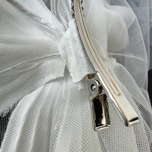 May include: A silver metal hair clip securing white tulle fabric. The clip has a curved design with a series of teeth along one edge. The tulle is layered and appears soft and delicate.
