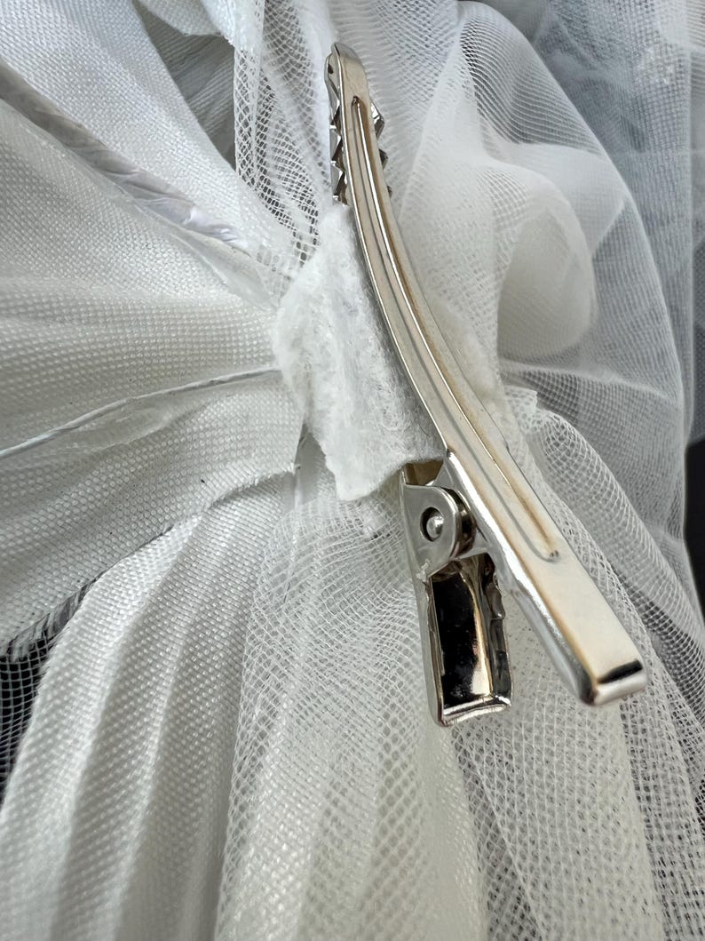 May include: A silver metal hair clip securing white tulle fabric. The clip has a curved design with a series of teeth along one edge. The tulle is layered and appears soft and delicate.