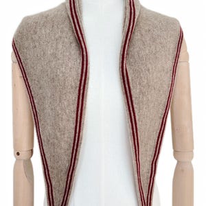 May include: A triangular knit scarf in a heathered beige color featuring a dark red and white striped border along the edges. The scarf is draped around the neck area, showcasing a textured knit pattern and pointed ends that hang down the front.