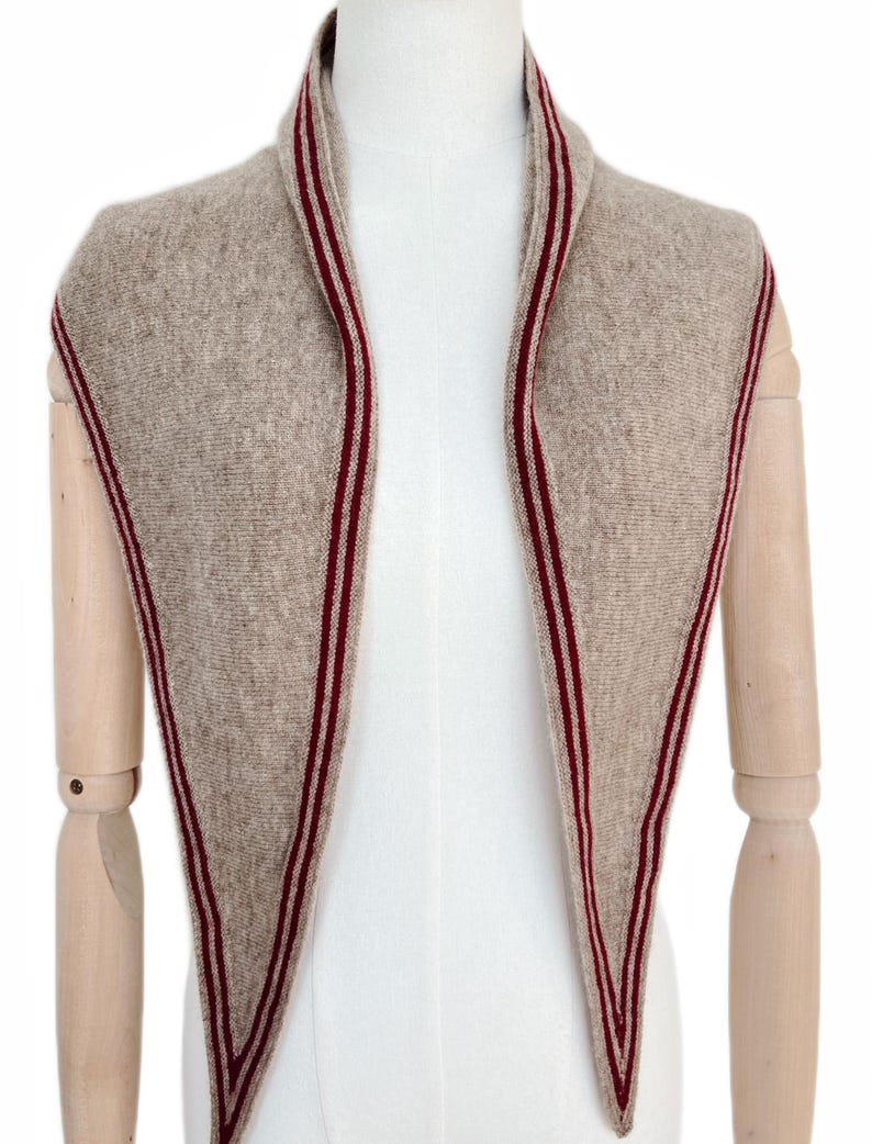 May include: A triangular knit scarf in a heathered beige color featuring a dark red and white striped border along the edges. The scarf is draped around the neck area, showcasing a textured knit pattern and pointed ends that hang down the front.