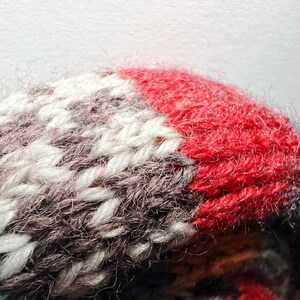 May include: Close-up of a knitted item with a textured surface. The item features a color block design with red, white, gray, and teal sections. The yarn appears soft and fluffy, with visible stitches.