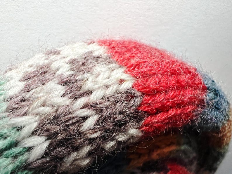 May include: Close-up of a knitted item with a textured surface. The item features a color block design with red, white, gray, and teal sections. The yarn appears soft and fluffy, with visible stitches.