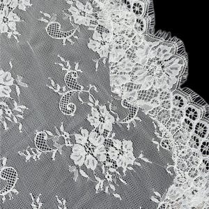 Op de afbeelding: Een witte kanten stof met een gedetailleerd bloemenpatroon, ingewikkeld borduurwerk en een geschulpte rand. De delicate mesh-achtergrond is versierd met herhalende bloemmotieven en bladranken, wat zorgt voor een textuurrijke en semi-transparante uitstraling.