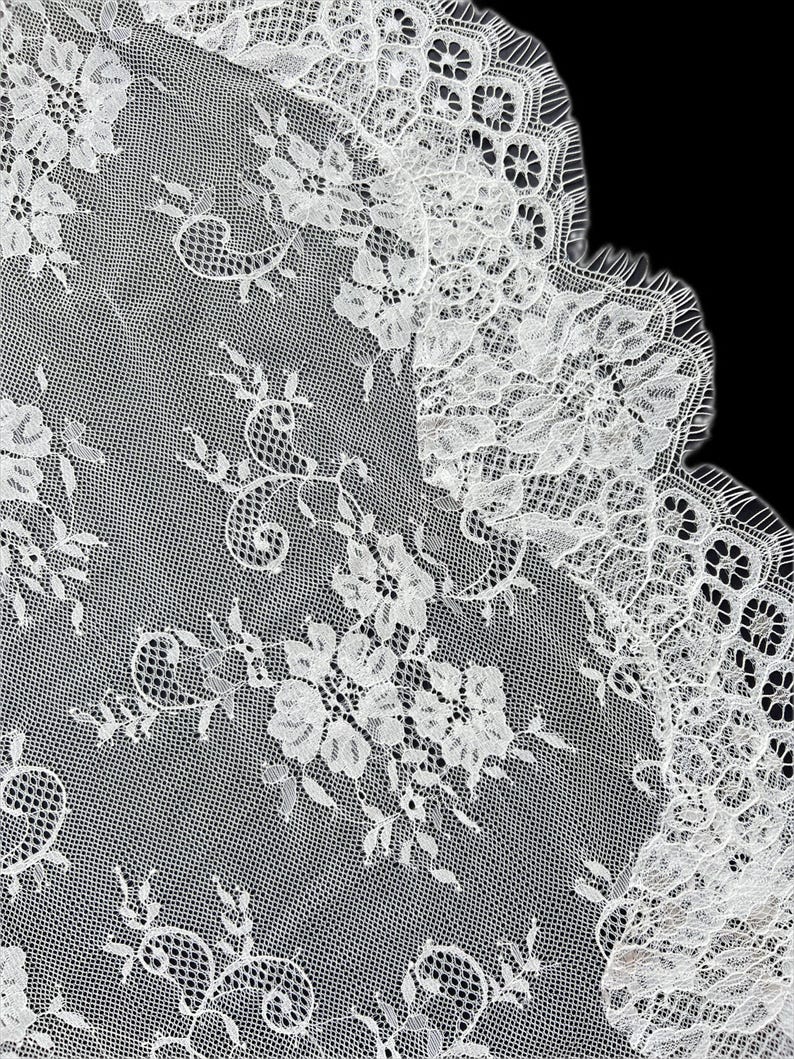 Op de afbeelding: Een witte kanten stof met een gedetailleerd bloemenpatroon, ingewikkeld borduurwerk en een geschulpte rand. De delicate mesh-achtergrond is versierd met herhalende bloemmotieven en bladranken, wat zorgt voor een textuurrijke en semi-transparante uitstraling.
