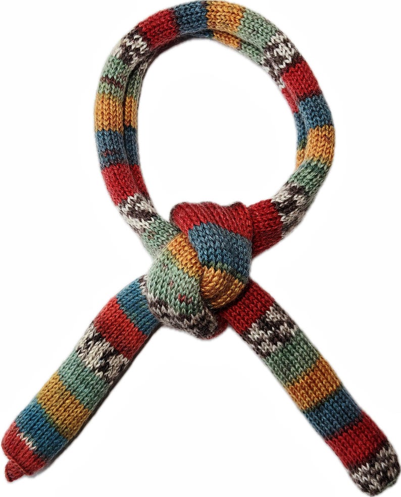 May include: A colorful, knitted scarf with a knotted design. The scarf features a striped pattern in shades of red, blue, green, yellow, and white. The ends of the scarf are tapered, and the overall design is eye-catching and unique.