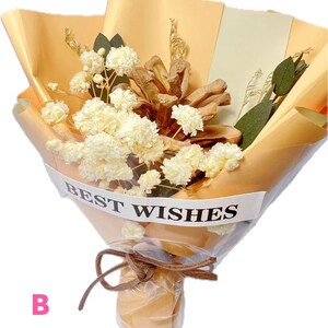 May include: A bouquet of dried white baby's breath flowers and a brown pinecone wrapped in peach-colored paper with a white band featuring the text BEST WISHES. The arrangement is tied with a brown cord and encased in clear plastic wrapping.