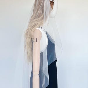 May include: A bridal veil with a floral headpiece. The veil is sheer and white, cascading down the back. The headpiece features white flowers and small pearl accents. The mannequin is wearing a black dress.