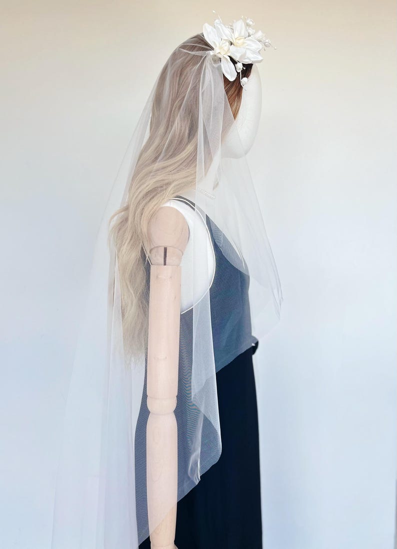 May include: A bridal veil with a floral headpiece. The veil is sheer and white, cascading down the back. The headpiece features white flowers and small pearl accents. The mannequin is wearing a black dress.
