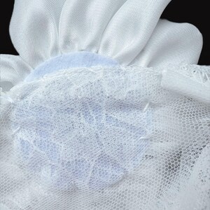 Op de afbeelding: Een wit stoffen bloemaccessoire met gladde, satijnachtige bloemblaadjes in een cirkelvormig patroon. De basis van de bloem is bevestigd aan een laag witte, fijnmazige netstof met een delicate kanten overlay met bloemmotief.