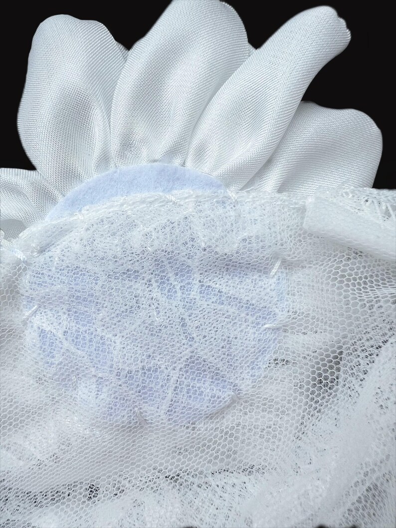 Op de afbeelding: Een wit stoffen bloemaccessoire met gladde, satijnachtige bloemblaadjes in een cirkelvormig patroon. De basis van de bloem is bevestigd aan een laag witte, fijnmazige netstof met een delicate kanten overlay met bloemmotief.