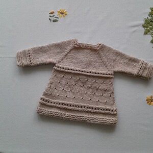 May include: A beige knitted baby dress with a delicate lace pattern. The dress has long sleeves and a round neckline.