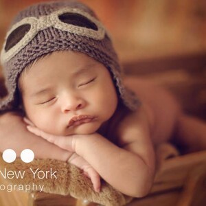 May include: A baby wearing a brown knitted aviator hat with a brown leather strap and goggles, sleeping with their arms crossed in front of them.