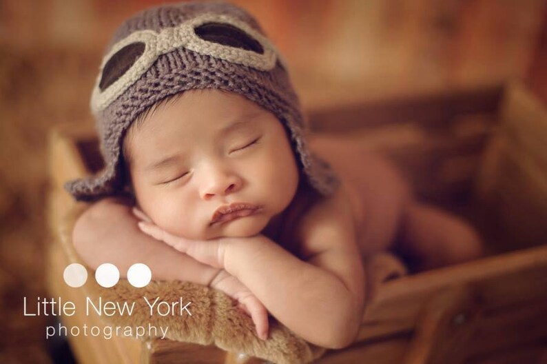 May include: A baby wearing a brown knitted aviator hat with a brown leather strap and goggles, sleeping with their arms crossed in front of them.