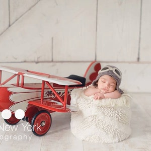 May include: A newborn baby wearing a gray aviator hat and goggles is sleeping in a white fluffy basket. A red toy airplane with white accents is in the background.