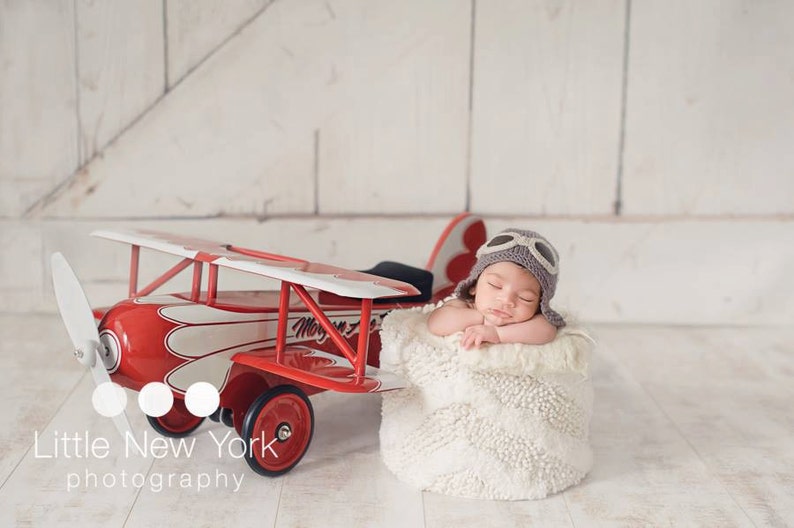 May include: A newborn baby wearing a gray aviator hat and goggles is sleeping in a white fluffy basket. A red toy airplane with white accents is in the background.