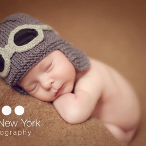 May include: A sleeping baby wearing a gray knitted hat with a brown leather aviator style goggles. The baby is lying on a brown blanket.