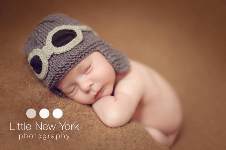 May include: A sleeping baby wearing a gray knitted hat with a brown leather aviator style goggles. The baby is lying on a brown blanket.