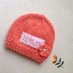 K&ouml;nnte beinhalten: Korallenfarbene gestrickte Baby-M&uuml;tze mit dem Namen "TEAGAN" auf einem wei&szlig;en Filzaufn&auml;her gestickt. Eine kleine Blume mit Perlenmitte ist angebracht. Die M&uuml;tze ist handgefertigt und f&uuml;r ein Neugeborenes geeignet.
