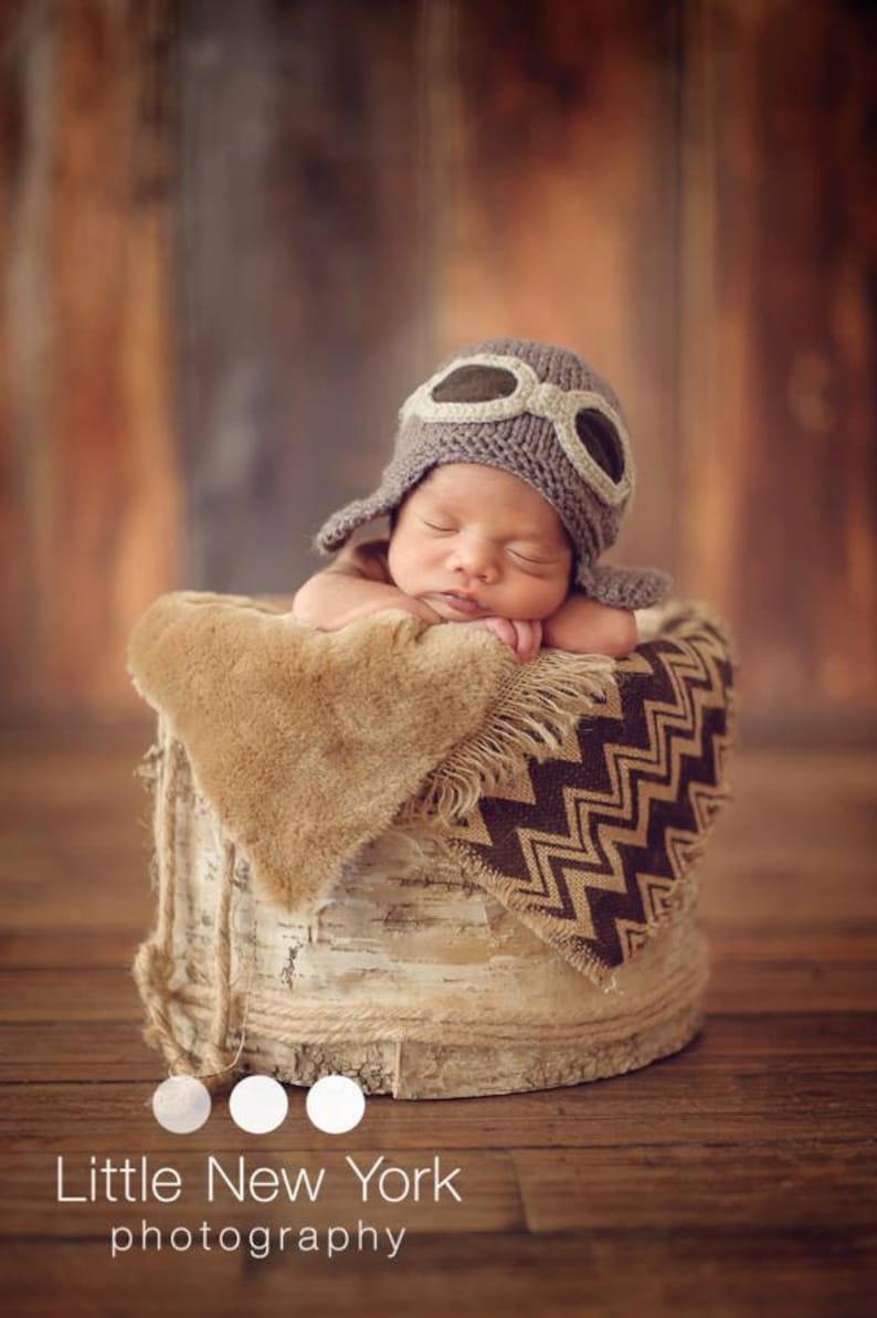 May include: A newborn baby wearing a gray knitted aviator hat with faux leather goggles sleeps in a birch bark bucket lined with a brown and tan chevron patterned blanket.