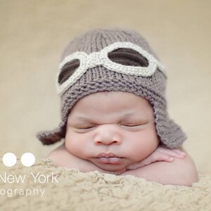 May include: A baby wearing a brown knitted aviator hat with white goggles. The baby is sleeping with their arms crossed in front of them.