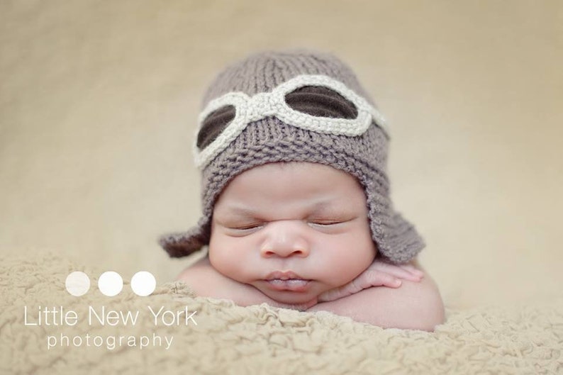 May include: A baby wearing a brown knitted aviator hat with white goggles. The baby is sleeping with their arms crossed in front of them.