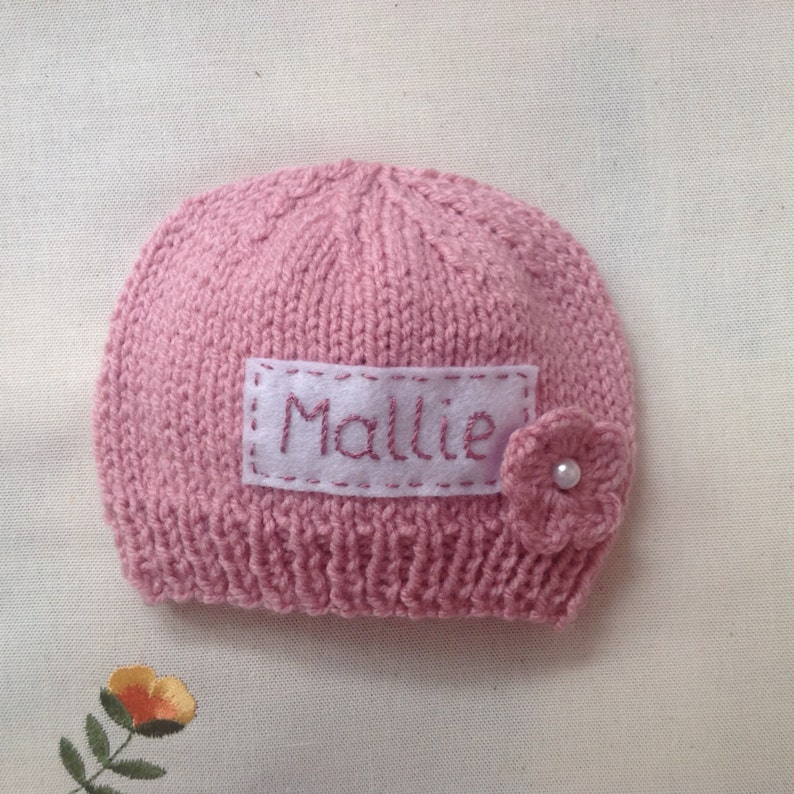K&ouml;nnte beinhalten: Ein rosa gestrickter Babyhut mit dem Namen "Mallie" auf einem wei&szlig;en Aufn&auml;her gestickt. Eine kleine rosa Blume mit Perlenmitte ist an der Seite befestigt. Der Hut ist aus weichem Garn gefertigt.