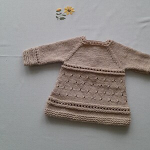 May include: A beige knitted baby dress with long sleeves and a delicate lace pattern.