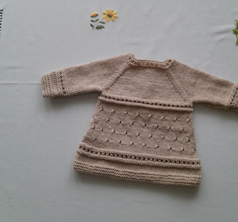 May include: A beige knitted baby dress with long sleeves and a delicate lace pattern.