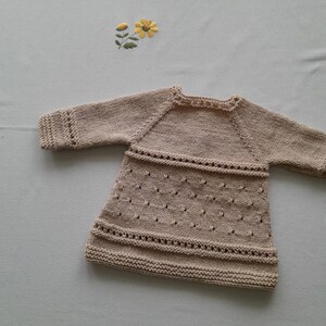 May include: A beige knitted baby dress with a delicate lace pattern. The dress has long sleeves and a round neckline.