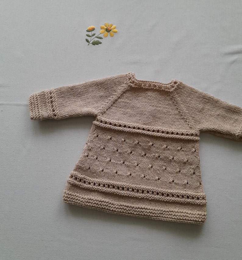 May include: A beige knitted baby dress with a delicate lace pattern. The dress has long sleeves and a round neckline.