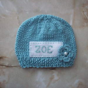 K&ouml;nnte beinhalten: Eine gestrickte t&uuml;rkisfarbene Baby-M&uuml;tze mit dem Namen "ZOE" auf einem wei&szlig;en Aufn&auml;her gestickt. Eine kleine Blume mit Perlenmitte ist an der Seite befestigt. Die M&uuml;tze ist ein weicher, handgefertigter Artikel.