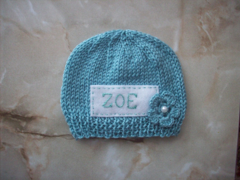 K&ouml;nnte beinhalten: Eine gestrickte t&uuml;rkisfarbene Baby-M&uuml;tze mit dem Namen "ZOE" auf einem wei&szlig;en Aufn&auml;her gestickt. Eine kleine Blume mit Perlenmitte ist an der Seite befestigt. Die M&uuml;tze ist ein weicher, handgefertigter Artikel.