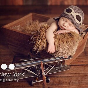 May include: A newborn baby wearing a gray knitted aviator hat with brown leather goggles sleeps in a wooden box lined with a brown faux fur blanket. A toy airplane sits in front of the box.