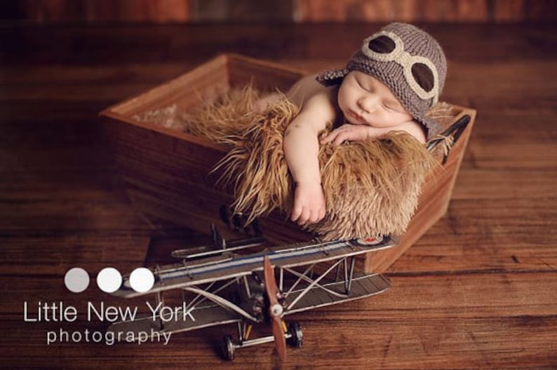 May include: A newborn baby wearing a gray knitted aviator hat with brown leather goggles sleeps in a wooden box lined with a brown faux fur blanket. A toy airplane sits in front of the box.