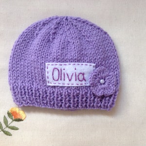 K&ouml;nnte beinhalten: Ein gestrickter, lavendelfarbener Babyhut mit dem Namen "Olivia" auf einem wei&szlig;en Aufn&auml;her gestickt. Eine kleine, geh&auml;kelte Blume mit Perlenmitte ist befestigt. Der Hut liegt auf einer cremefarbenen Oberfl&auml;che.