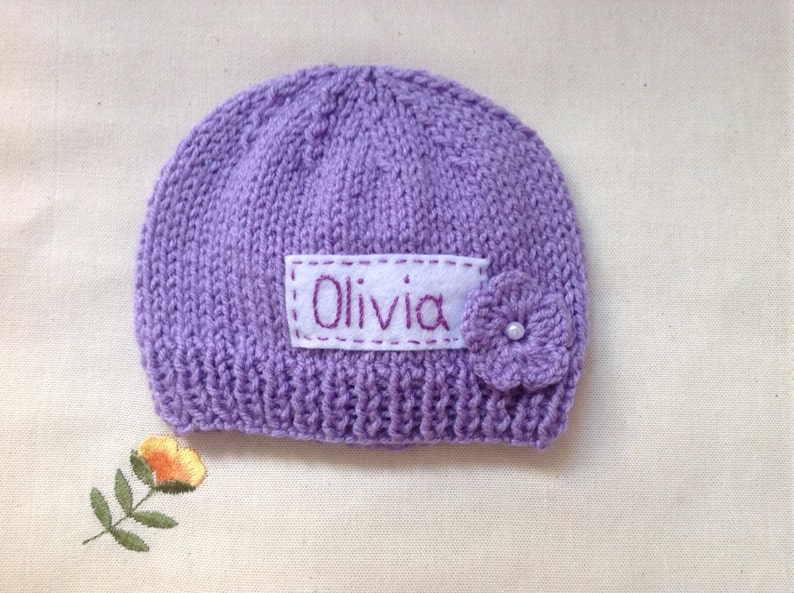 K&ouml;nnte beinhalten: Ein gestrickter, lavendelfarbener Babyhut mit dem Namen "Olivia" auf einem wei&szlig;en Aufn&auml;her gestickt. Eine kleine, geh&auml;kelte Blume mit Perlenmitte ist befestigt. Der Hut liegt auf einer cremefarbenen Oberfl&auml;che.