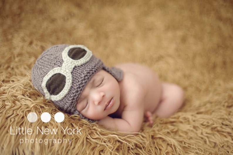 May include: A baby wearing a gray knitted hat with white aviator goggles sleeps on a brown fuzzy blanket.